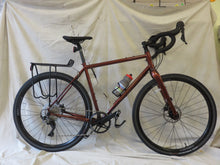 Load image into Gallery viewer, A Jamis Renegade S2 gravel bicycle with a steel frame, brown color, and equipped with a rear rack and tubeless tires, displayed on a white background.
